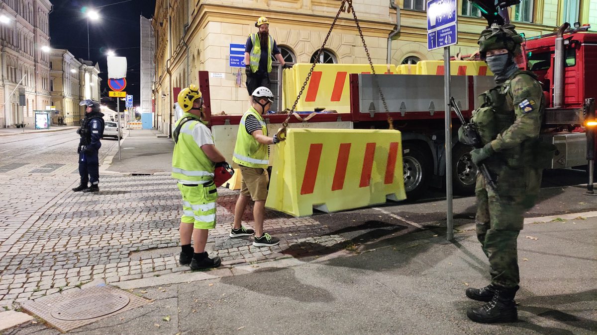 Kolme staralaista valmiusharjoituksessa öisessä Helsingissä. Miehet siirtävät betoniporsaita autosta kadun sulkemiseksi. Poliisi ja sotilas partioivat vieressä.